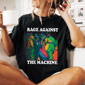 May include: Black t-shirt with the text "RAGE AGAINST THE MACHINE" in white. The graphic depicts a frog-like figure facing a line of police officers. The frog wears a blue scarf. The image has a colorful, comic book style.