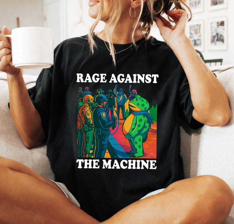 May include: Black t-shirt with the text "RAGE AGAINST THE MACHINE" in white. The graphic depicts a frog-like figure facing a line of police officers. The frog wears a blue scarf. The image has a colorful, comic book style.