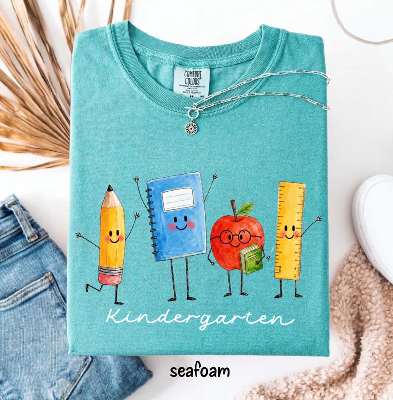 May include: A teal crewneck t-shirt featuring a graphic of a smiling pencil, a blue spiral notebook, a red apple wearing glasses, and a yellow ruler. The text "Kindergarten" is printed in white script below the characters. A silver chain necklace rests on the collar.