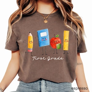 May include: A dark brown short-sleeve t-shirt featuring a graphic of a yellow pencil, a blue notebook, a red apple with glasses, and a yellow ruler, all with smiling faces. The text "First Grade" is printed in white below the characters.