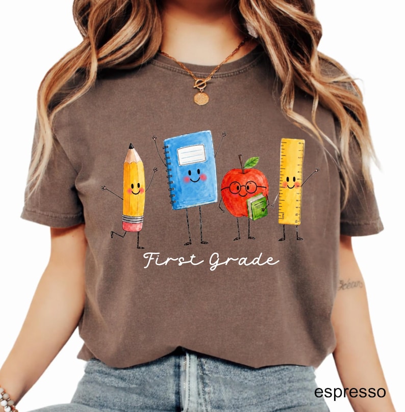 May include: A dark brown short-sleeve t-shirt featuring a graphic of a yellow pencil, a blue notebook, a red apple with glasses, and a yellow ruler, all with smiling faces. The text "First Grade" is printed in white below the characters.