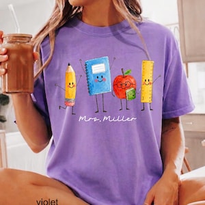 May include: A purple t-shirt featuring a graphic of a yellow pencil, a blue notebook, a red apple with glasses, and a yellow ruler, all with smiling faces. The text "Mrs. Miller" is printed in white script below the school supply characters.
