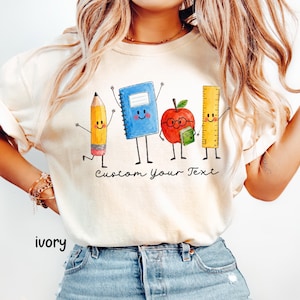 May include: An ivory t-shirt featuring a watercolor-style graphic of four anthropomorphic school supplies: a yellow pencil, a blue spiral notebook, a red apple with glasses holding a small book, and a yellow ruler. The text "Custom Your Text" is printed below.