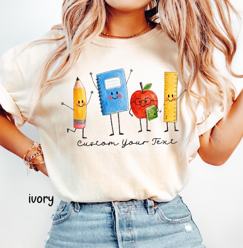 May include: An ivory t-shirt featuring a watercolor-style graphic of four anthropomorphic school supplies: a yellow pencil, a blue spiral notebook, a red apple with glasses holding a small book, and a yellow ruler. The text "Custom Your Text" is printed below.