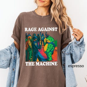 May include: Espresso-colored t-shirt with the text "RAGE AGAINST THE MACHINE" in white. The graphic features a cartoon frog and figures in police uniforms. The shirt has rolled-up sleeves and a relaxed fit.
