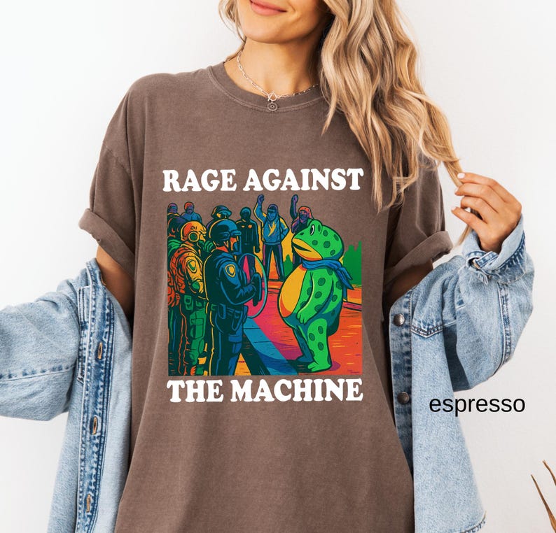 May include: Espresso-colored t-shirt with the text "RAGE AGAINST THE MACHINE" in white. The graphic features a cartoon frog and figures in police uniforms. The shirt has rolled-up sleeves and a relaxed fit.