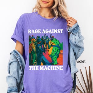 May include: Violet t-shirt with the text "RAGE AGAINST THE MACHINE" in white block letters. The graphic depicts a green frog facing a group of police officers. The frog wears a blue scarf. The shirt is a casual style.