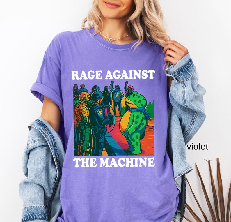 May include: Violet t-shirt with the text "RAGE AGAINST THE MACHINE" in white block letters. The graphic depicts a green frog facing a group of police officers. The frog wears a blue scarf. The shirt is a casual style.