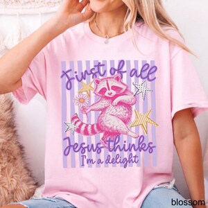 May include: A light pink t-shirt featuring a cartoon raccoon holding a flower, with the text "First of all Jesus thinks I'm a delight." The design includes purple stripes, gold stars, and a daisy.