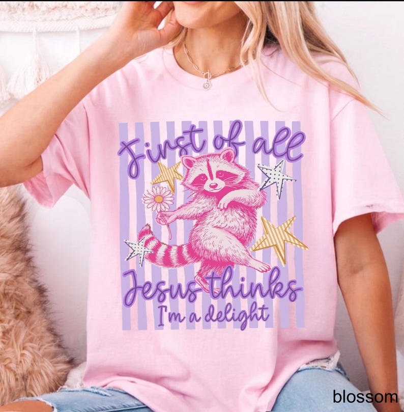 May include: A light pink t-shirt featuring a cartoon raccoon holding a flower, with the text "First of all Jesus thinks I'm a delight." The design includes purple stripes, gold stars, and a daisy.