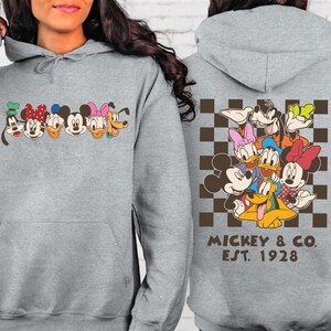 Puede incluir: Una sudadera con capucha gris claro con una l&iacute;nea de caras de personajes de dibujos animados en el pecho. La parte posterior de la sudadera con capucha muestra un grupo de personajes sobre un fondo a cuadros marr&oacute;n y negro, con el texto "MICKEY & CO. EST. 1928".