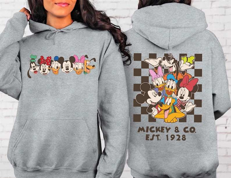 Puede incluir: Una sudadera con capucha gris claro con una l&iacute;nea de caras de personajes de dibujos animados en el pecho. La parte posterior de la sudadera con capucha muestra un grupo de personajes sobre un fondo a cuadros marr&oacute;n y negro, con el texto "MICKEY & CO. EST. 1928".