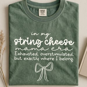 May include: Moss green t-shirt with white text: "in my string cheese mama era. Exhausted, overstimulated, but exactly where I belong." A white bow graphic is below the text. The word "moss" is at the bottom.