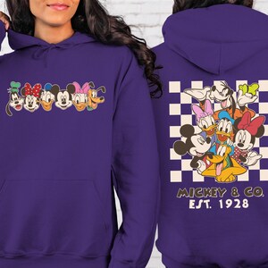 Puede incluir: Una sudadera con capucha morada con caras de personajes de dibujos animados en la parte delantera y trasera. La parte delantera muestra caras de personajes, mientras que la parte trasera presenta un grupo de personajes sobre un fondo a cuadros, con el texto "Mickey & Co. Est. 1928".
