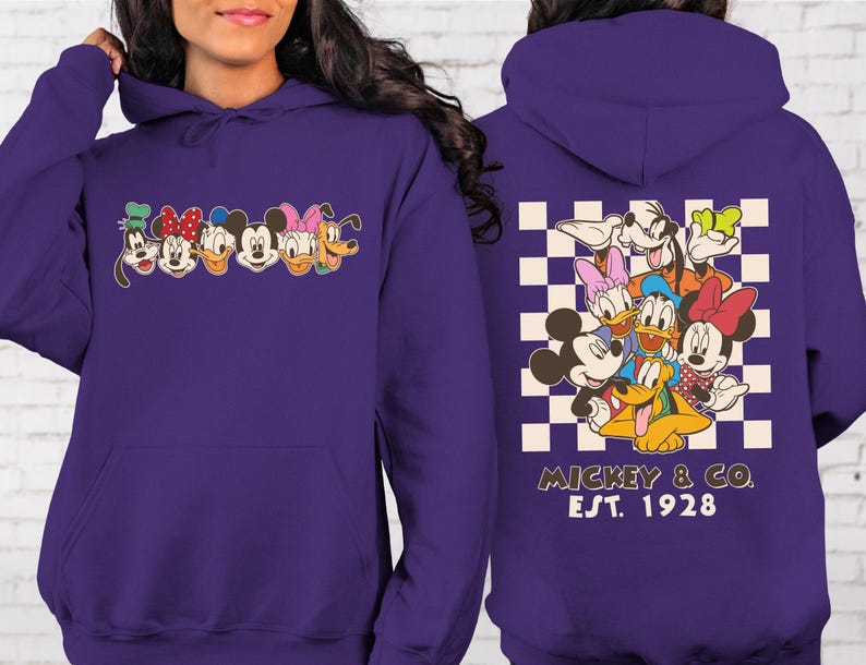Puede incluir: Una sudadera con capucha morada con caras de personajes de dibujos animados en la parte delantera y trasera. La parte delantera muestra caras de personajes, mientras que la parte trasera presenta un grupo de personajes sobre un fondo a cuadros, con el texto "Mickey & Co. Est. 1928".