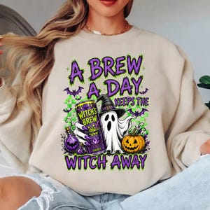 May include: A beige crewneck sweatshirt featuring a graphic print with the text A BREW A DAY KEEPS THE WITCH AWAY. The design includes a ghost wearing a witch hat, a can labeled WITCH'S BREW, a pumpkin, and purple bats on a green checkered background.