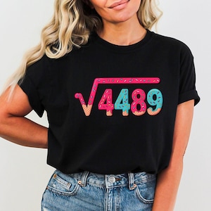 May include: Black t-shirt featuring a colorful graphic of a square root symbol with the number 4489 inside, designed to look like a donut. The graphic includes pink, blue, and tan colors. The shirt is made of a soft material.