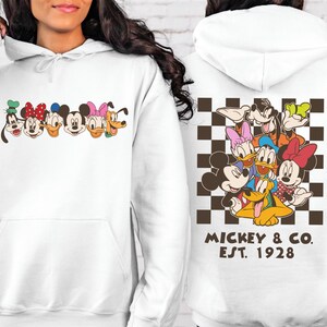 Puede incluir: Sudadera con capucha blanca con un gr&aacute;fico de personajes de dibujos animados en la parte delantera. La parte trasera presenta un grupo de personajes sobre un fondo a cuadros marr&oacute;n y blanco, con el texto "Mickey & Co. Est. 1928".