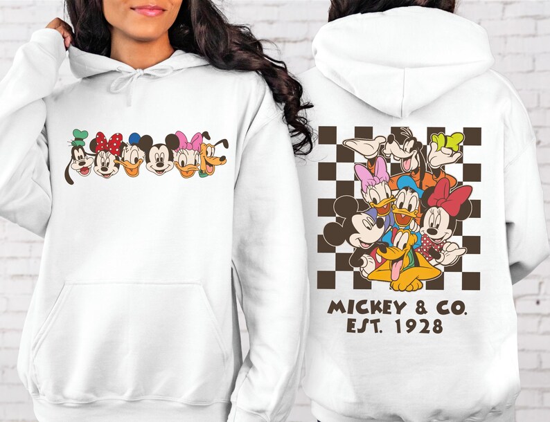 Puede incluir: Sudadera con capucha blanca con un gr&aacute;fico de personajes de dibujos animados en la parte delantera. La parte trasera presenta un grupo de personajes sobre un fondo a cuadros marr&oacute;n y blanco, con el texto "Mickey & Co. Est. 1928".