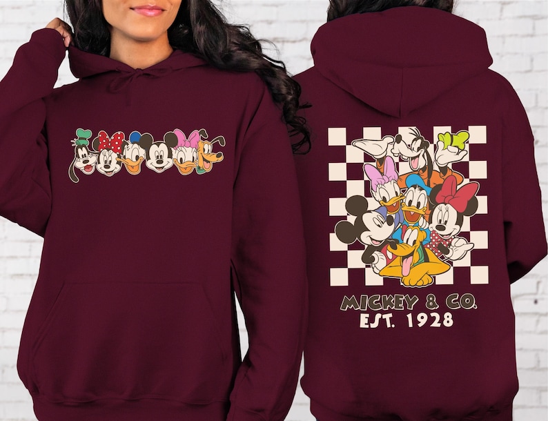 Puede incluir: Sudadera con capucha burdeos con gr&aacute;ficos de personajes de dibujos animados en la parte delantera y trasera. La parte delantera muestra caras de personajes, y la parte trasera presenta un grupo de personajes sobre un fondo a cuadros. El texto "Mickey & Co. Est. 1928" est&aacute; impreso debajo.