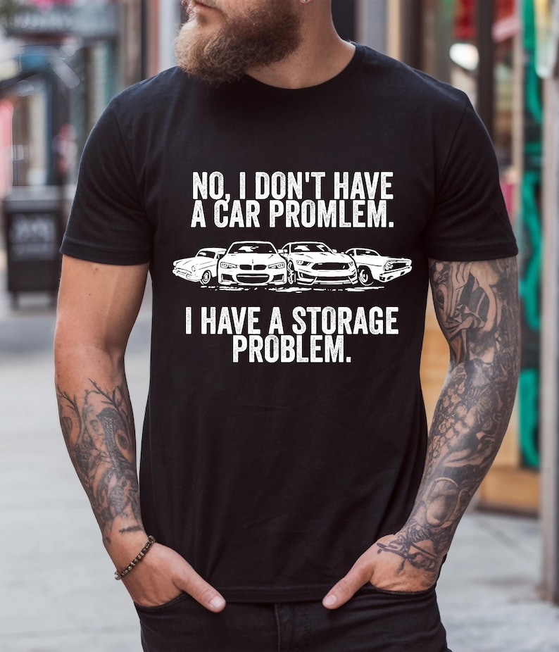 May include: Black t-shirt with white text: "NO, I DON'T HAVE A CAR PROMLEM. I HAVE A STORAGE PROBLEM." Below the text are illustrations of four cars. The shirt is a crew neck and has short sleeves.