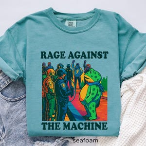 May include: Seafoam green t-shirt with the text "RAGE AGAINST THE MACHINE" above a colorful graphic. The graphic depicts a frog facing a line of police officers. The shirt is made of Comfort Colors material.
