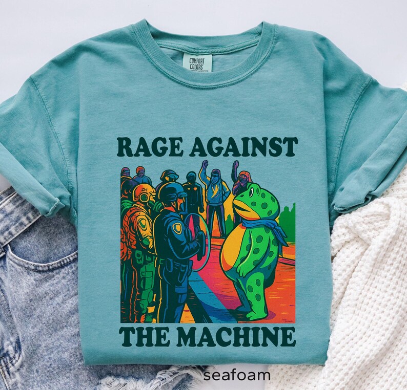 May include: Seafoam green t-shirt with the text "RAGE AGAINST THE MACHINE" above a colorful graphic. The graphic depicts a frog facing a line of police officers. The shirt is made of Comfort Colors material.