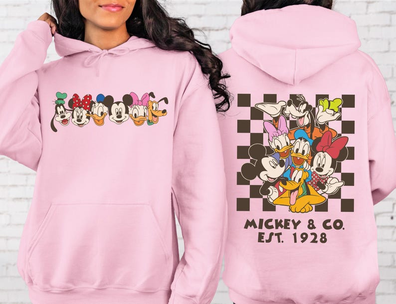 Puede incluir: Una sudadera con capucha rosa claro con un dise&ntilde;o de dibujos animados de Mickey Mouse y sus amigos. La parte delantera muestra las caras de los personajes, mientras que la parte trasera muestra un grupo de personajes sobre un fondo a cuadros marr&oacute;n y blanco con "Mickey & Co. Est. 1928."