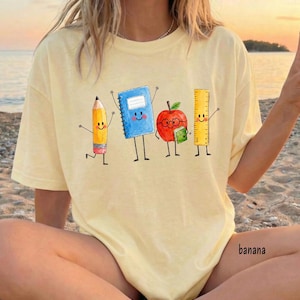 May include: A light yellow t-shirt featuring a graphic of four anthropomorphic school supplies: a yellow pencil, a blue spiral notebook, a red apple with glasses holding a small green book, and a yellow ruler, all with smiling faces.