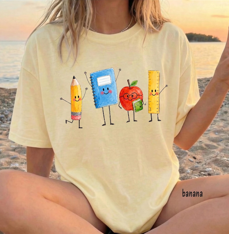May include: A light yellow t-shirt featuring a graphic of four anthropomorphic school supplies: a yellow pencil, a blue spiral notebook, a red apple with glasses holding a small green book, and a yellow ruler, all with smiling faces.
