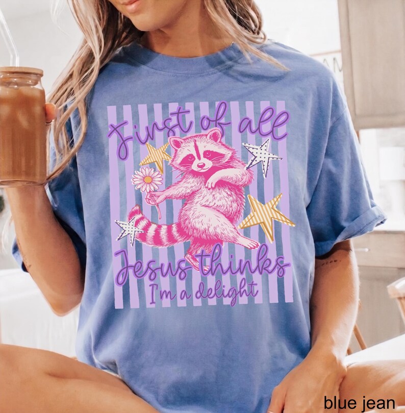 May include: Blue jean-colored t-shirt featuring a pink raccoon holding a daisy, with text that reads "First of all Jesus thinks I'm a delight." The design includes stars and vertical stripes.
