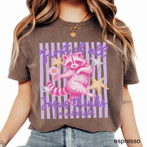May include: Espresso-colored t-shirt featuring a pink raccoon holding a flower, surrounded by stars. The text reads "First of all Jesus thinks I'm a delight" in a purple script. The design is set against a backdrop of purple stripes.