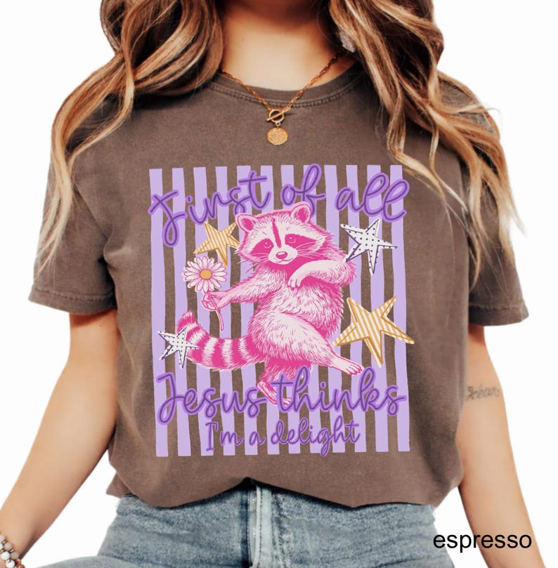 May include: Espresso-colored t-shirt featuring a pink raccoon holding a flower, surrounded by stars. The text reads "First of all Jesus thinks I'm a delight" in a purple script. The design is set against a backdrop of purple stripes.