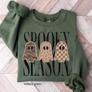 May include: A military green sweatshirt featuring the black text SPOOKY SEASON. Below the text are three ghost shapes with different patterns: one with a camouflage print, one with a leopard print, and one with a brown and tan checkerboard pattern.