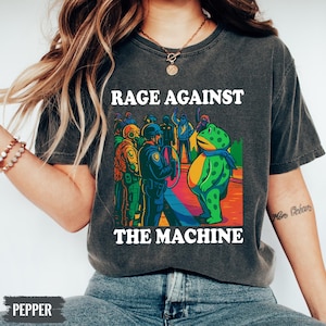 May include: Dark gray t-shirt with the text "RAGE AGAINST THE MACHINE" in white. The graphic features a cartoon frog facing a group of people in riot gear. The frog wears a blue scarf. The image has a vintage, distressed look.