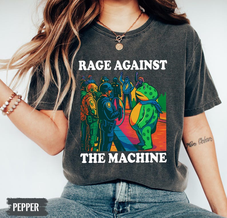 May include: Dark gray t-shirt with the text "RAGE AGAINST THE MACHINE" in white. The graphic features a cartoon frog facing a group of people in riot gear. The frog wears a blue scarf. The image has a vintage, distressed look.