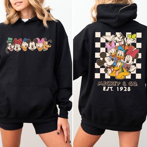 Puede incluir: Sudadera con capucha negra con un dise&ntilde;o de personajes de Disney. La parte delantera muestra caras de personajes, mientras que la parte trasera presenta un grupo de personajes sobre un fondo a cuadros con "Mickey & Co. Est. 1928".