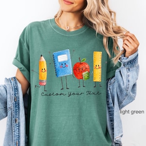 May include: A light green t-shirt featuring a graphic of a smiling pencil, a blue notebook, a red apple wearing glasses, and a yellow ruler. Below the characters, the text Custom Your Text is printed in a black, cursive, handwritten-style font.