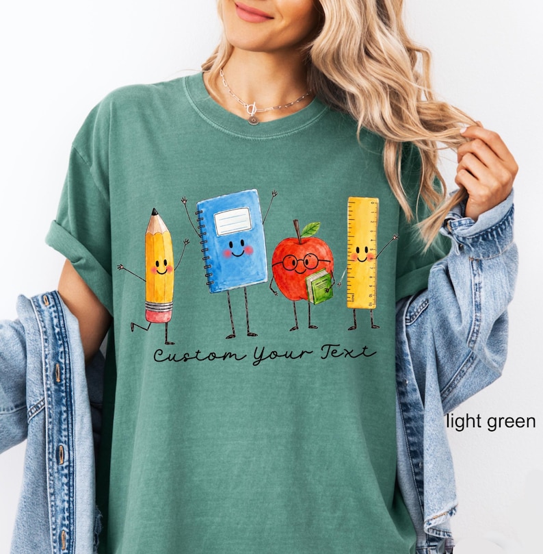 May include: A light green t-shirt featuring a graphic of a smiling pencil, a blue notebook, a red apple wearing glasses, and a yellow ruler. Below the characters, the text Custom Your Text is printed in a black, cursive, handwritten-style font.