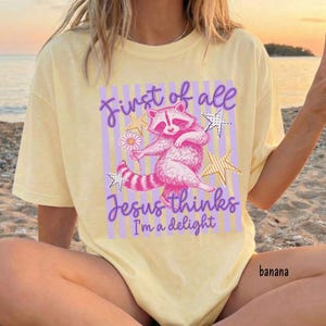 May include: A light yellow t-shirt with a graphic featuring a pink raccoon holding a flower, surrounded by stars. The text reads "First of all Jesus thinks I'm a delight." The word "banana" is printed on the bottom right.