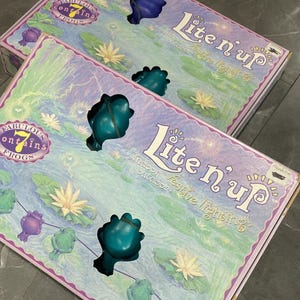 May include: Two boxes of "Lite n' up" indoor/outdoor festive lighting. The boxes feature a design with lily pads, frogs, and the text "Fabulous contains 7 frogs". The boxes are purple and green.