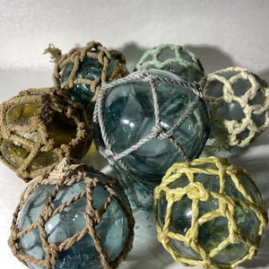Puede incluir: Colección de siete flotadores de pesca de vidrio en varios tonos de verde y amarillo, envueltos en redes de cuerda anudada. Los flotadores varían en tamaño y color, creando una estética rústica y náutica.