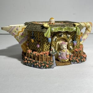 May include: A decorative teapot-shaped figurine with a cat figure in a doorway. The teapot has a brown thatched roof, floral accents, and a handle with a floral pattern. The base is surrounded by orange roses. The text "Happy Easter" is visible.