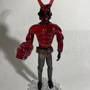 May include: A red glass figurine of a horned demon with a muscular physique. The figure has a black face and is wearing gray pants and a belt. It stands on a clear, textured base.