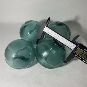 Puede incluir: Se muestran cuatro esferas de vidrio color turquesa, cada una con un diámetro de aproximadamente 8 cm. Un calibrador digital mide el diámetro de una esfera. Las esferas tienen una forma ligeramente irregular y una calidad translúcida.