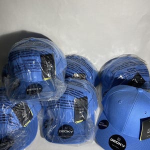 Puede incluir: Un conjunto de gorras de béisbol azules, algunas aún en su embalaje de plástico original. Cada gorra presenta una visera curva y el logotipo "DECKY". Las gorras son de un color azul sólido y vibrante. La imagen está bien iluminada.