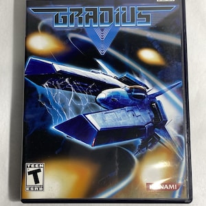 以下が含まれることがあります： PlayStation 2のゲームソフト「グラディウス」のケース。カバーには、暗い青と黒の背景に、稲妻のエフェクトを持つ青い宇宙船が描かれています。ゲームはESRBによってTeen（13歳以上対象）にレーティングされています。