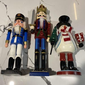 Peut inclure: Trois casse-noisettes de Noël en bois. L'un est un soldat en uniforme bleu, un autre est un roi avec une couronne et une épée, et le troisième est un bonhomme de neige tenant un arbre et un cadeau. Les casse-noisettes sont peints de couleurs vives.