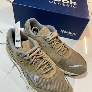 May include: A pair of Reebok Classic sneakers in tan and gray, with mesh and suede details. The shoes are new, with a tag attached, and are displayed next to a blue Reebok Classic box.