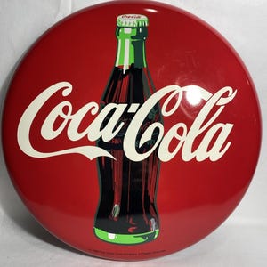 Puede incluir: Un letrero redondo y rojo de Coca-Cola con el logotipo de Coca-Cola en escritura blanca. Un gráfico de botella verde y negra está en el centro. El letrero tiene un acabado brillante y probablemente está hecho de metal.